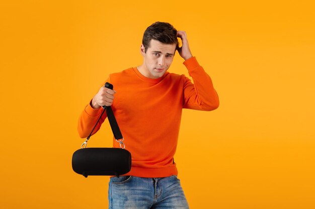 Élégant jeune homme souriant en pull orange tenant un haut-parleur sans fil heureux d'écouter de la musique s'amuser style coloré bonne humeur isolé sur fond jaune