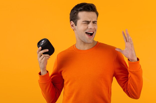 Élégant jeune homme souriant en pull orange tenant haut-parleur sans fil heureux d'écouter de la musique