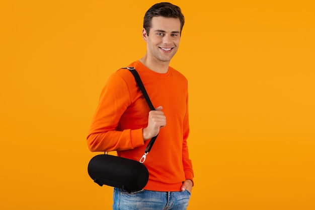 Élégant jeune homme souriant en pull orange tenant haut-parleur sans fil heureux d'écouter de la musique