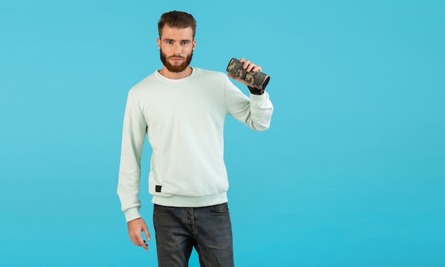 Élégant jeune homme souriant tenant haut-parleur sans fil, écouter de la musique sur fond bleu