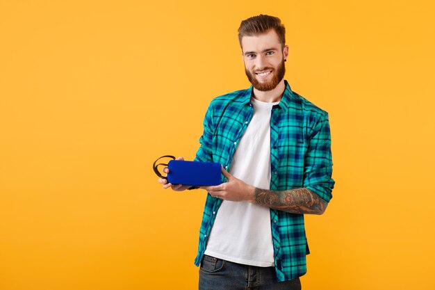 Élégant jeune homme souriant tenant haut-parleur sans fil heureux d'écouter de la musique style coloré de bonne humeur isolé sur mur jaune