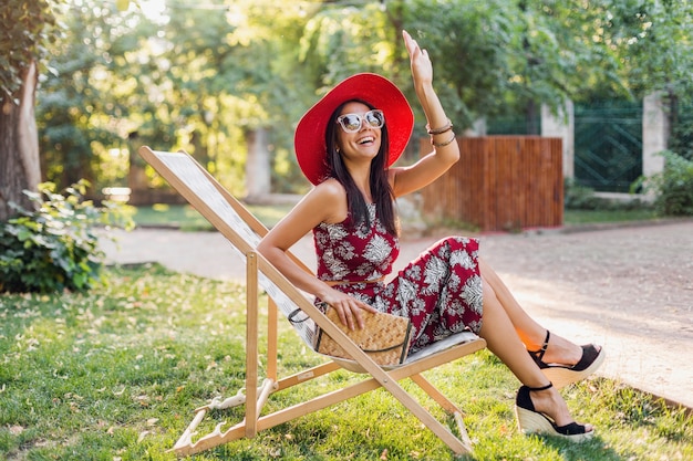 Élégante belle femme assise dans une chaise longue en tenue de style tropical, agitant la main, tendance de la mode estivale, sac à main en paille, chapeau rouge, lunettes de soleil, accessoires, souriant, bonne humeur, vacances