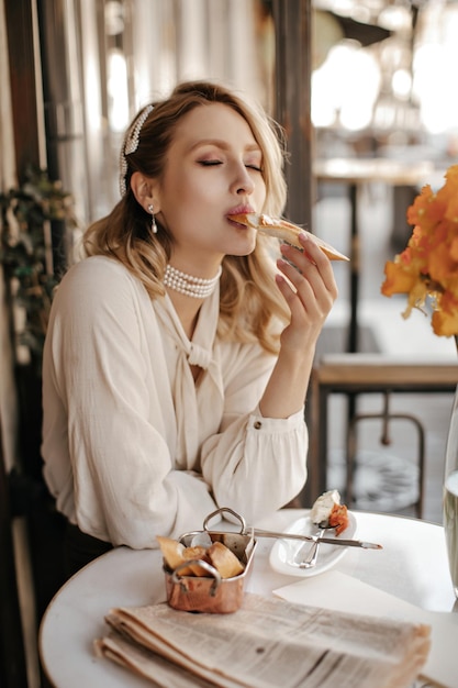 Élégante belle femme blonde en collier de perles et chemisier léger est assise dans un café et savoure une délicieuse bruschetta