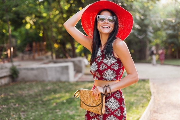 Élégante belle femme marchant dans le parc en tenue tropicale. dame dans la tendance de la mode estivale de style rue. portant un sac à main en paille, un chapeau rouge, des lunettes de soleil, des accessoires. fille souriante de bonne humeur en vacances.