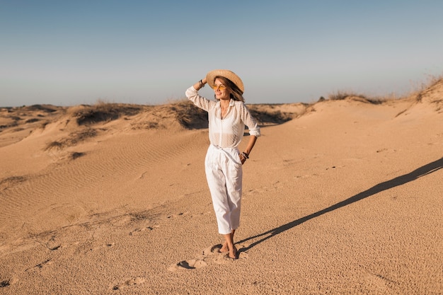 Élégante belle femme marchant dans le sable du désert en tenue blanche portant un chapeau de paille sur le coucher du soleil