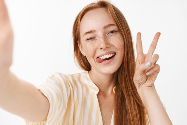 Élégante belle femme rousse avec de jolies taches de rousseur sortant la langue et souriant de joie en un clin d'œil montrant joyeusement signe de paix, prenant selfie sur smartphone