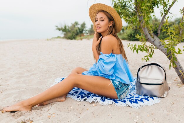 Élégante belle femme souriante assise sur le sable avec des jambes maigres en vacances d'été sur la plage tropicale portant un chapeau de paille, sac à dos argenté