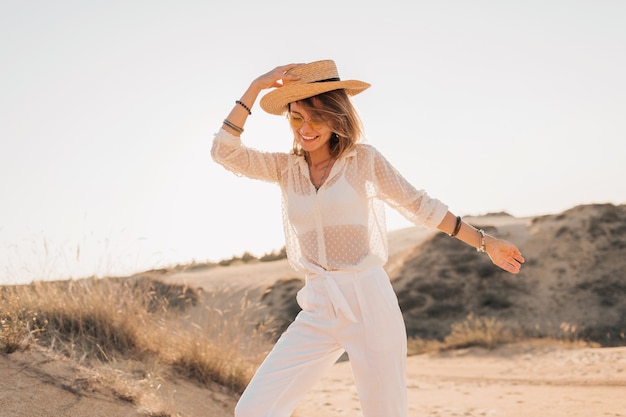 Élégante belle femme souriante heureuse posant dans le sable du désert en tenue blanche portant un chapeau de paille et des lunettes de soleil sur le coucher du soleil