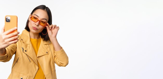 Élégante belle fille asiatique à lunettes de soleil prenant selfie sur smartphone posant pour une photo tenant un téléphone mobile fond de studio blanc