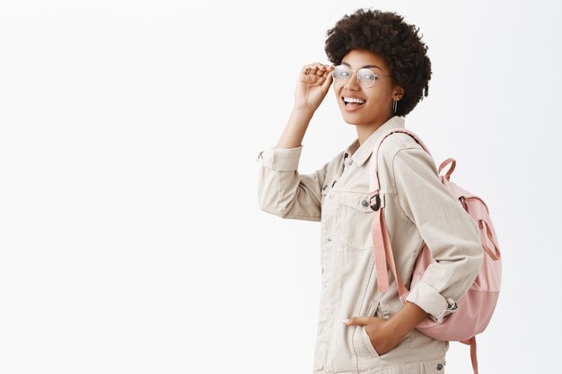 Élégante belle touriste avec une peau foncée et une coiffure afro vérifiant les lunettes sur les yeux et tournant en se tenant debout de profil sur un mur gris