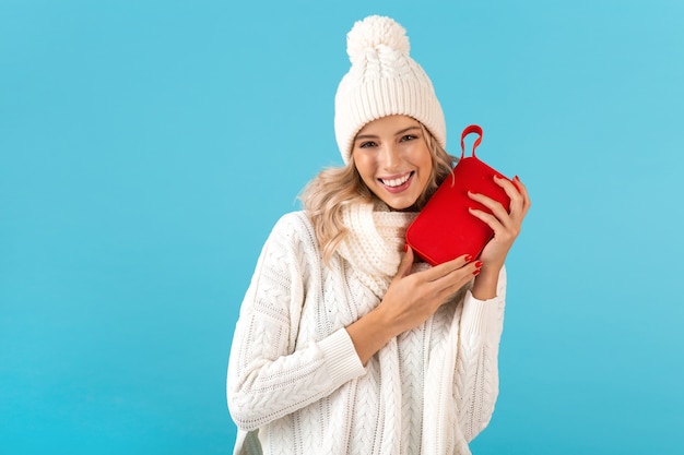 Élégante blonde souriante belle jeune femme tenant haut-parleur sans fil écouter de la musique heureux portant chandail blanc et bonnet tricoté posant sur bleu