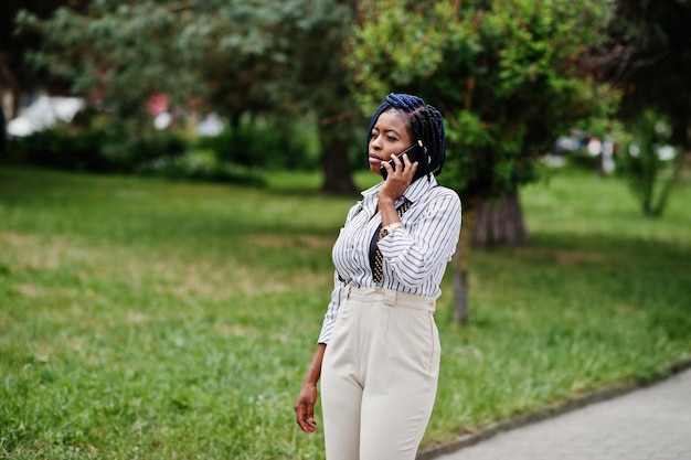 Élégante femme d'affaires afro-américaine au pantalon avec bretelles et chemisier posé en plein air et parlant au téléphone mobile
