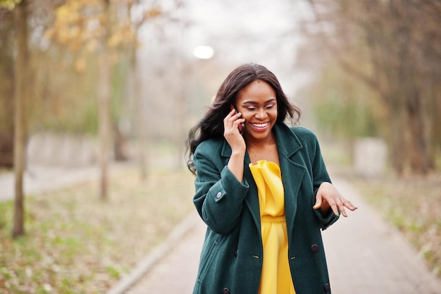 Élégante femme afro-américaine au manteau vert et à la robe jaune posée contre le parc d'automne et parlant au téléphone