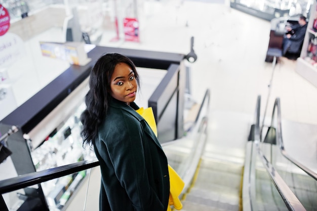 Élégante femme afro-américaine au manteau vert et robe jaune posée à l'escalator du centre commercial