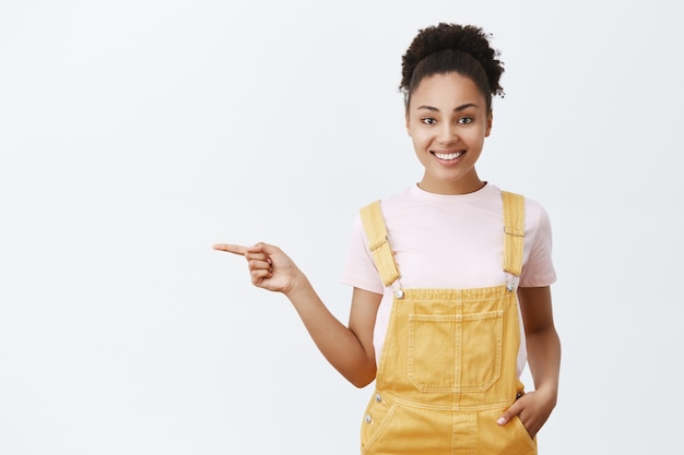 Élégante femme afro-américaine polie et amicale en salopette jaune, pointant vers la gauche et souriant largement, donnant des directions