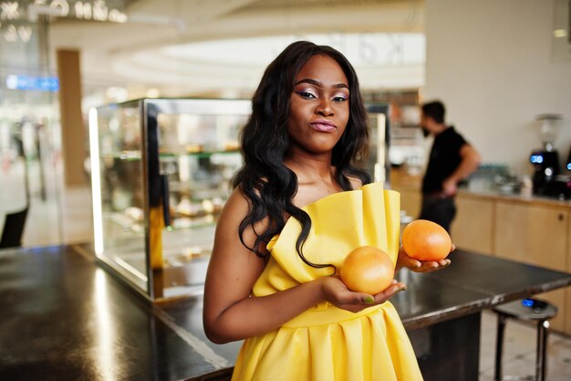 Élégante femme afro-américaine à la robe jaune posée au café avec deux pamplemousses sur les mains