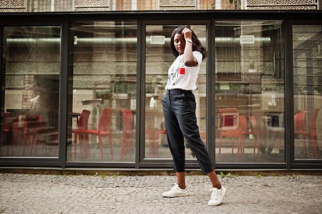 Élégante femme afro-américaine en t-shirt imprimé posé en plein air contre la fenêtre