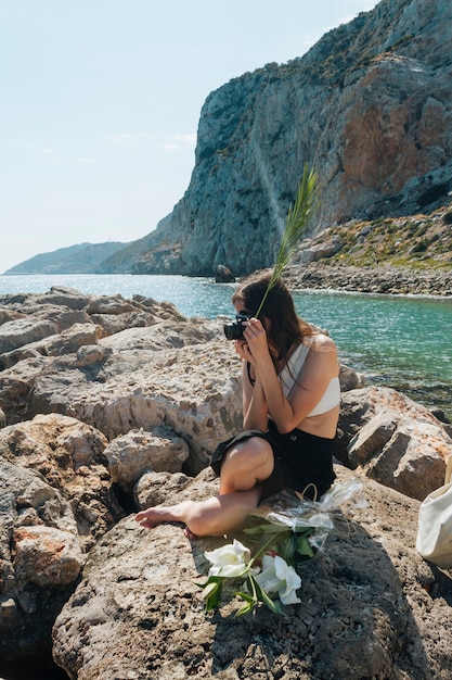 Élégante femme assise sur un rocher tenant des feuilles de palmier tout en prenant une photo avec l'appareil photo