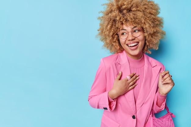 Élégante femme aux cheveux bouclés vêtue d'une élégante veste rose porte des sourires de sac regarde loin heureux d'entendre un compliment isolé sur fond bleu espace vide pour votre contenu publicitaire