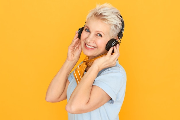 Élégante femme blonde à la retraite appréciant la musique, écoutant la radio dans des écouteurs sans fil posant contre jaune