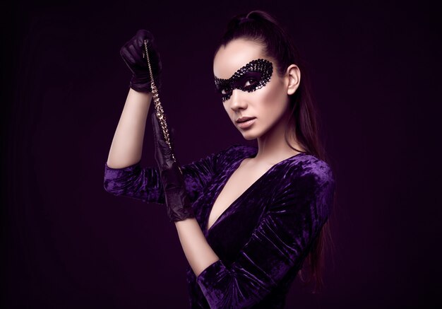 Élégante femme brune en masque de paillettes avec des gants noirs se penche sur le collier de diamants