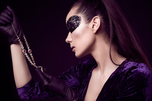 Élégante femme brune en masque de paillettes avec des gants noirs se penche sur le collier de diamants