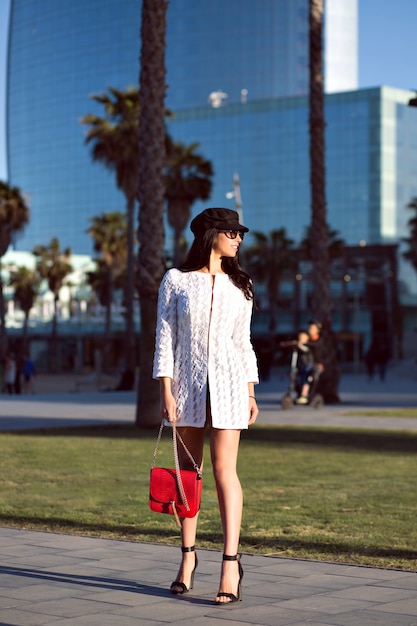 Élégante femme brune qui marche, tenue de style luxe fantaisie, bâtiments modernes et palmiers, couleurs toniques.