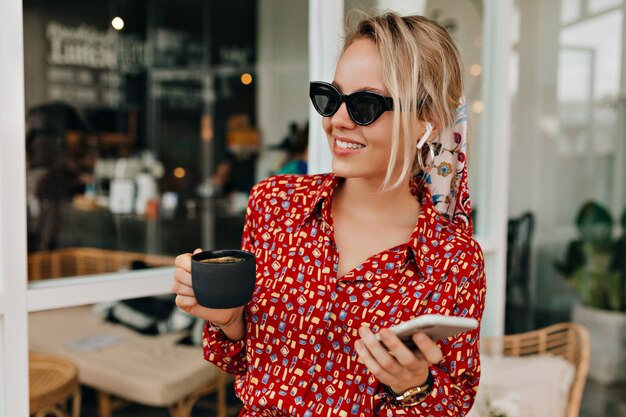 Élégante femme élégante portant des lunettes de soleil noires et une robe moderne lumineuse avec une tasse de café