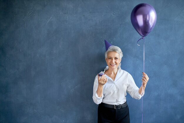 Élégante femme mature aux cheveux gris portant un chapeau de cône et des vêtements élégants mangeant un macaron à la fête, tenant un ballon à air