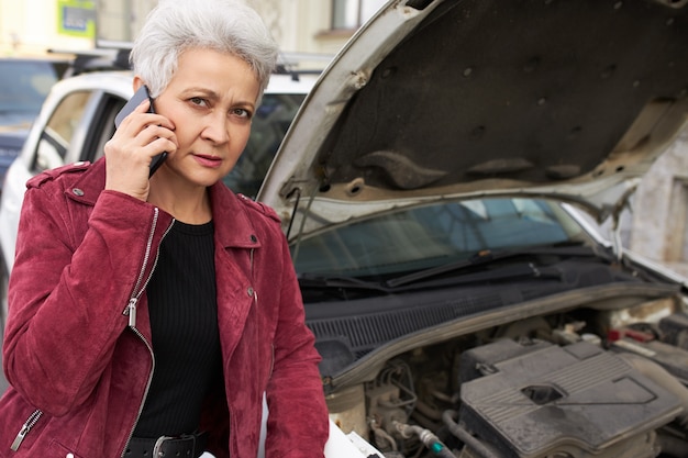 Élégante femme mûre aux cheveux gris attrayante debout près de sa voiture blanche cassée avec capot ouvert et parler au téléphone