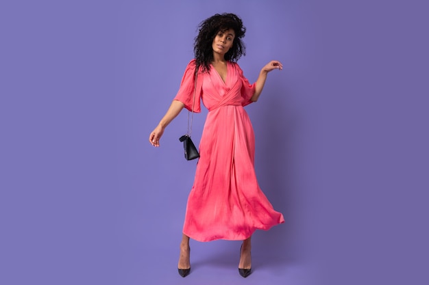 Élégante femme noire en robe de soirée rose s'amusant sur un mur violet. Porter des talons. Longueur totale.