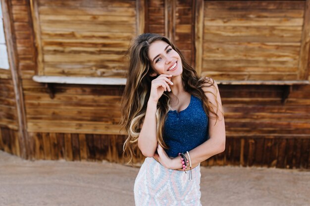 Élégante fille aux cheveux longs dans de jolis bracelets souriant joyeusement, tout en posant devant un bâtiment à colombages