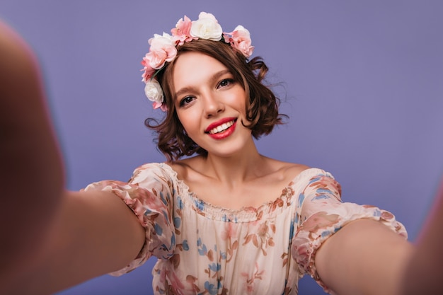 Élégante fille blanche dans un cercle de fleurs faisant selfie et souriant. Portrait de femme inspirée détendue avec coupe de cheveux courte.