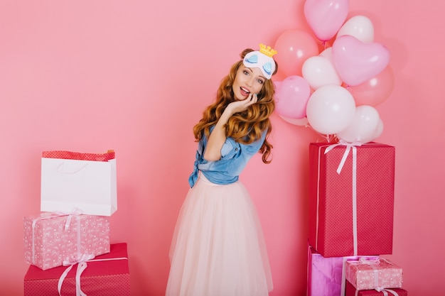 Élégante fille bouclée en jupe tendance luxuriante est surprise par le nombre de cadeaux pour son anniversaire d'amis. Charmante jeune femme aux cheveux longs en masque de sommeil posant avec des cadeaux et des ballons d'hélium à la fête