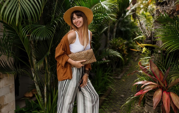 Élégante fille élégante en haut blanc et chapeau de paille posant sur des feuilles de palmier à Bali.