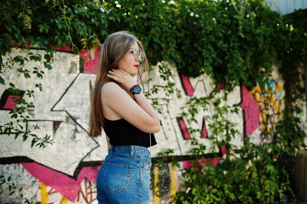 Élégante fille hipster décontractée dans des lunettes de casquette et des jeans porter de la musique d'écoute à partir d'écouteurs de téléphone portable contre un grand mur de graffitis