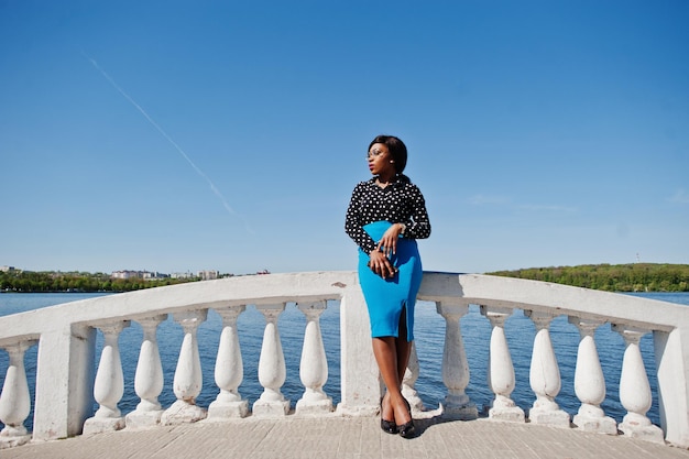 Élégante fille modèle afro-américaine en jupe bleue à lunettes et chemisier noir posé en plein air sur le pont contre le lac