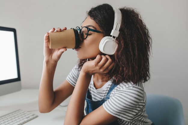 Élégante fille noire avec une coiffure frisée, boire du café sur le lieu de travail posant près de mur gris