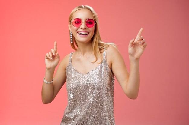 Élégante et insouciante fabuleuse jeune femme blonde en robe argentée avec des lunettes de soleil rouges s'amusant à danser joyeusement une discothèque sur une piste de danse souriant largement en profitant de passer du temps entre amis, fond de studio debout.
