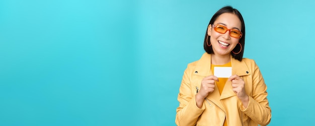 Élégante jeune femme asiatique à lunettes de soleil montrant une carte de crédit et souriante recommandant un paiement bancaire sans contact ou des réductions en magasin debout sur fond bleu