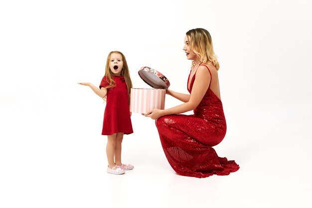 Élégante jeune femme blonde en robe rouge assis sur le sol avec boîte, lui donnant un cadeau d'anniversaire adorable fille