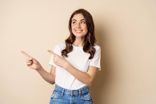 Élégante jeune femme caucasienne souriante pointant les doigts vers la gauche montrant une offre promotionnelle publicitaire standi...