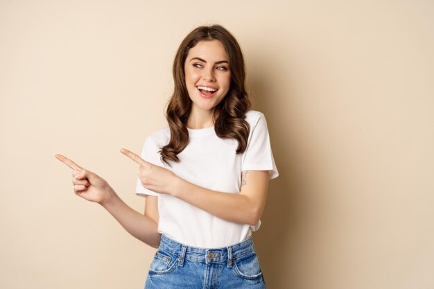 Élégante jeune femme caucasienne souriante, pointant les doigts vers la gauche, montrant une publicité, une offre promotionnelle, debout sur fond beige.