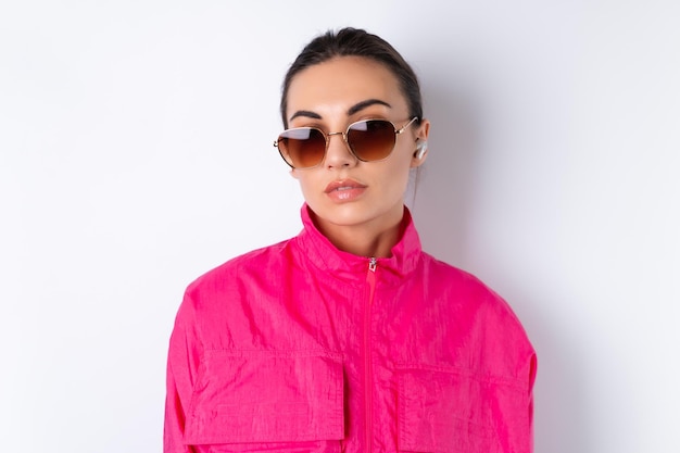 Élégante jeune femme dans une veste de sport rose vif, des lunettes de soleil à la mode et des écouteurs sans fil dans ses oreilles sur fond blanc