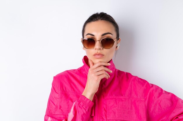 Élégante jeune femme dans une veste de sport rose vif, des lunettes de soleil à la mode et des écouteurs sans fil dans ses oreilles sur fond blanc