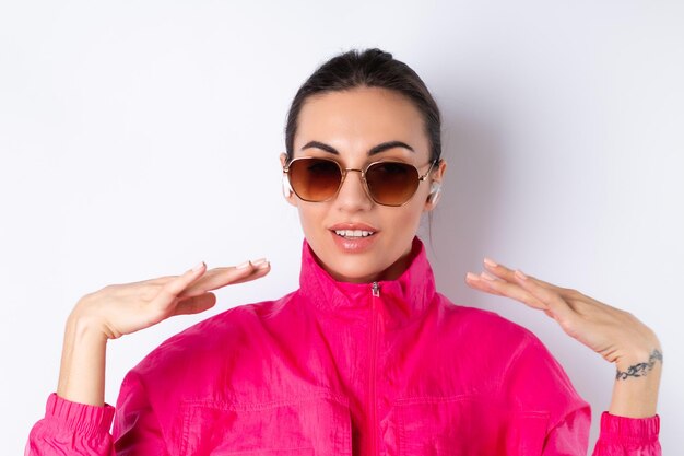 Élégante jeune femme dans une veste de sport rose vif, des lunettes de soleil à la mode et des écouteurs sans fil dans ses oreilles sur fond blanc