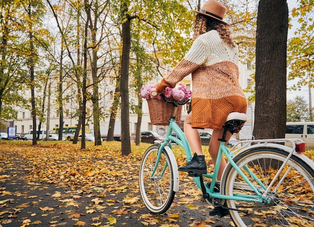 Élégante jeune femme faisant du vélo avec des fleurs dans la rue d'automne