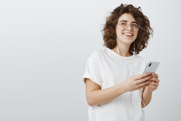 Élégante jeune femme à lunettes faisant une liste de lecture dans le téléphone, écouter de la musique via des écouteurs