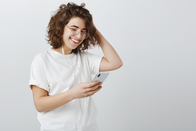 Élégante jeune femme à lunettes faisant une liste de lecture dans le téléphone, écouter de la musique via des écouteurs