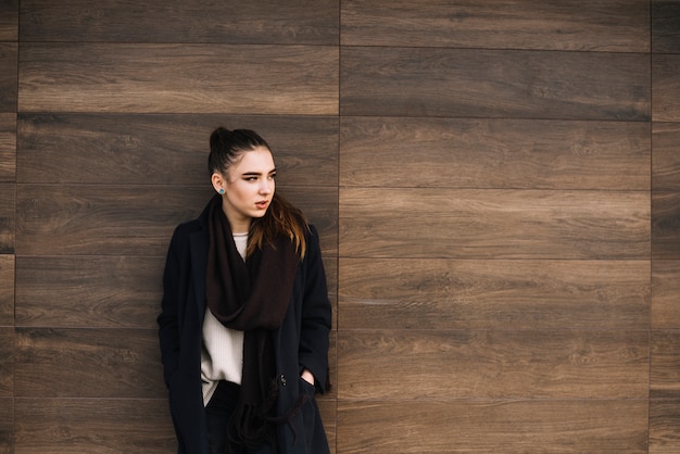 Élégante jeune femme en manteau avec écharpe près du mur en bois